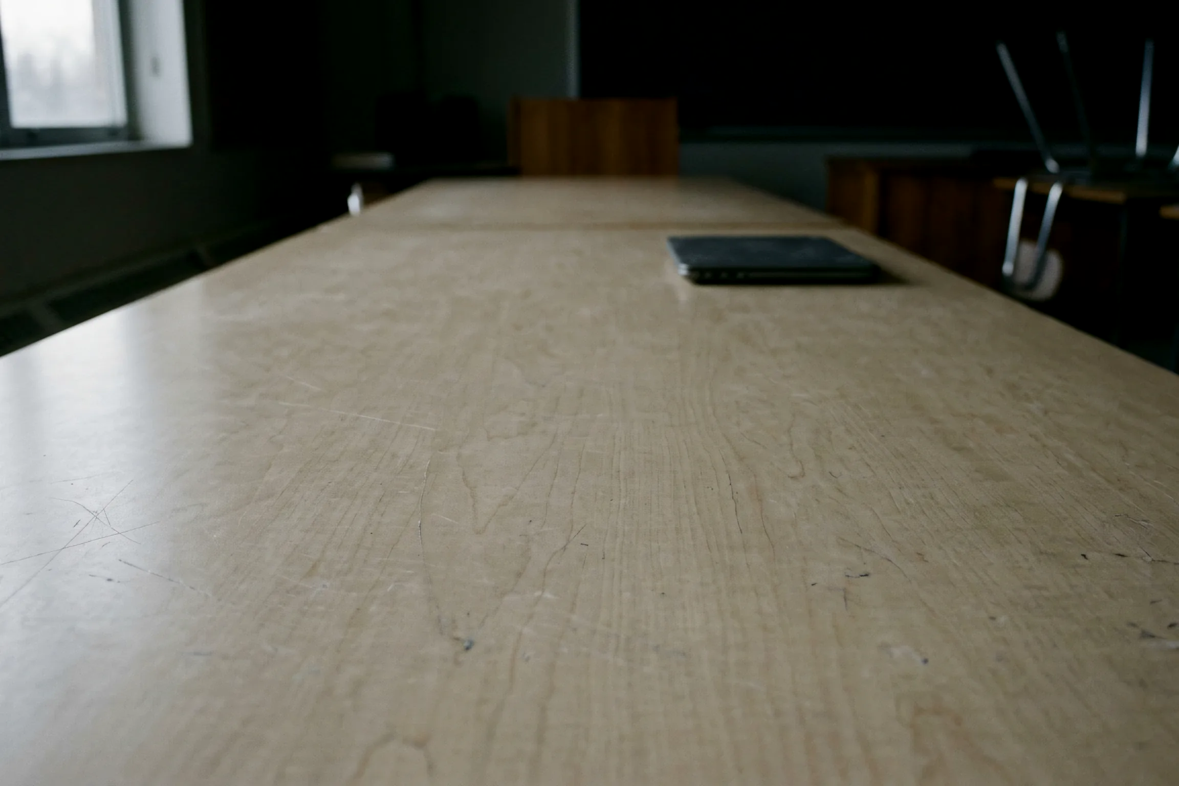 A closed, unbranded laptop alone on a wide empty laminate table, lit only by flat gray daylight from off frame.