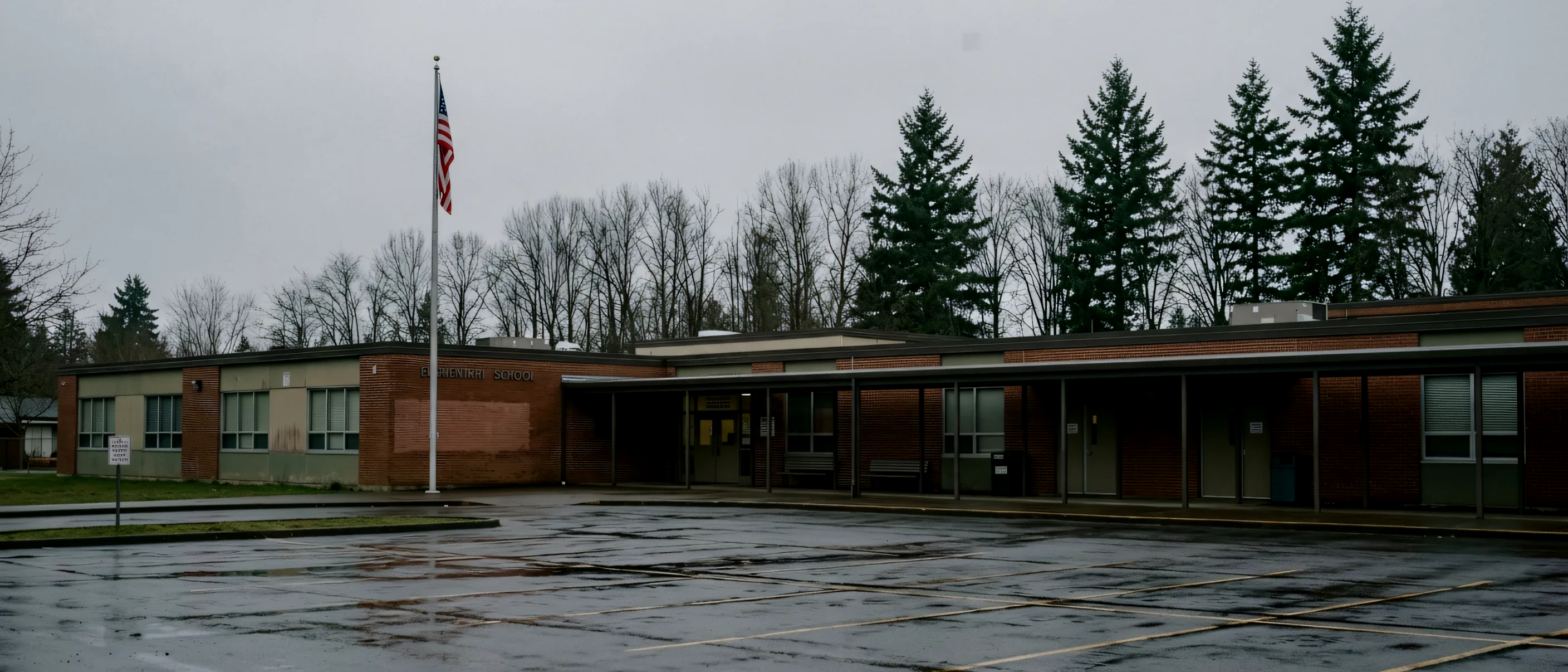 A single-storey public elementary school across a wet parking lot on an overcast morning, bare trees and firs behind.